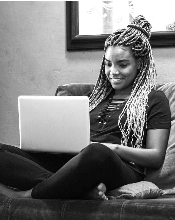 Young black woman with pigtails using notebook sitting on blue sofa in home room